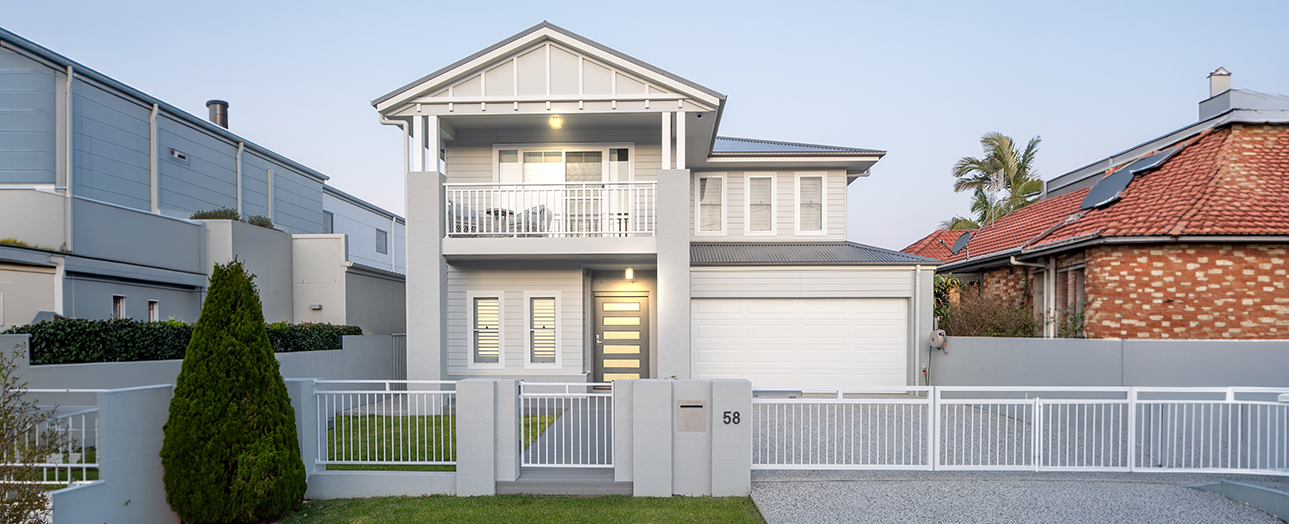 Modern two-story residential home in New Lambton with white facade, horizontal stripe detailing on front door, second-story balcony, white metal fencing, and integrated garage. House number 58 displayed on front gate. Manicured lawn with conical shrub in foreground. Neighboring homes visible on either side under clear sky.