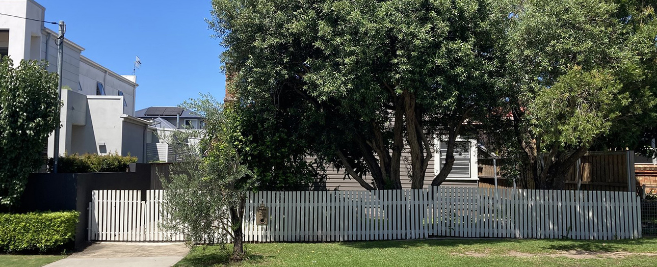 Residential street in New Lambton showing a weathered two-story house with white and gray exterior, surrounded by mature trees and a white picket fence, under clear blue sky.