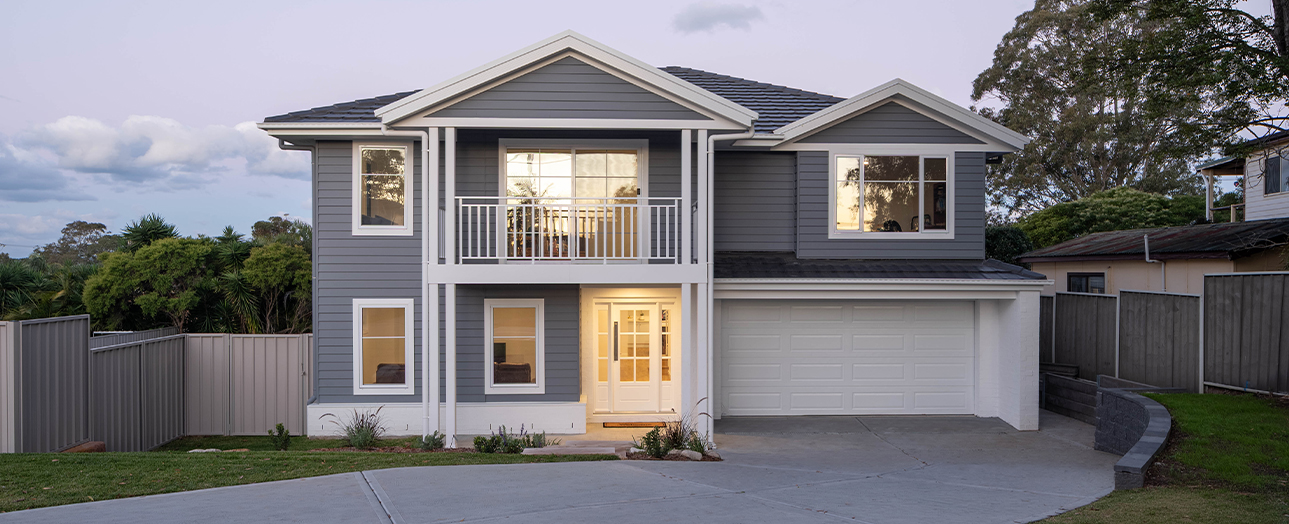 Modern two-story gray and white house with illuminated windows, white garage door, front balcony, and manicured lawn bordered by metal fencing, set in a residential neighborhood at dusk.