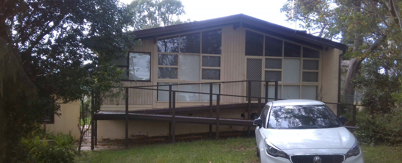 A single-story elevated house with cream and dark corrugated metal exterior, large windows, and a metal railing deck, surrounded by mature trees. A white sedan is parked in front on the grass.