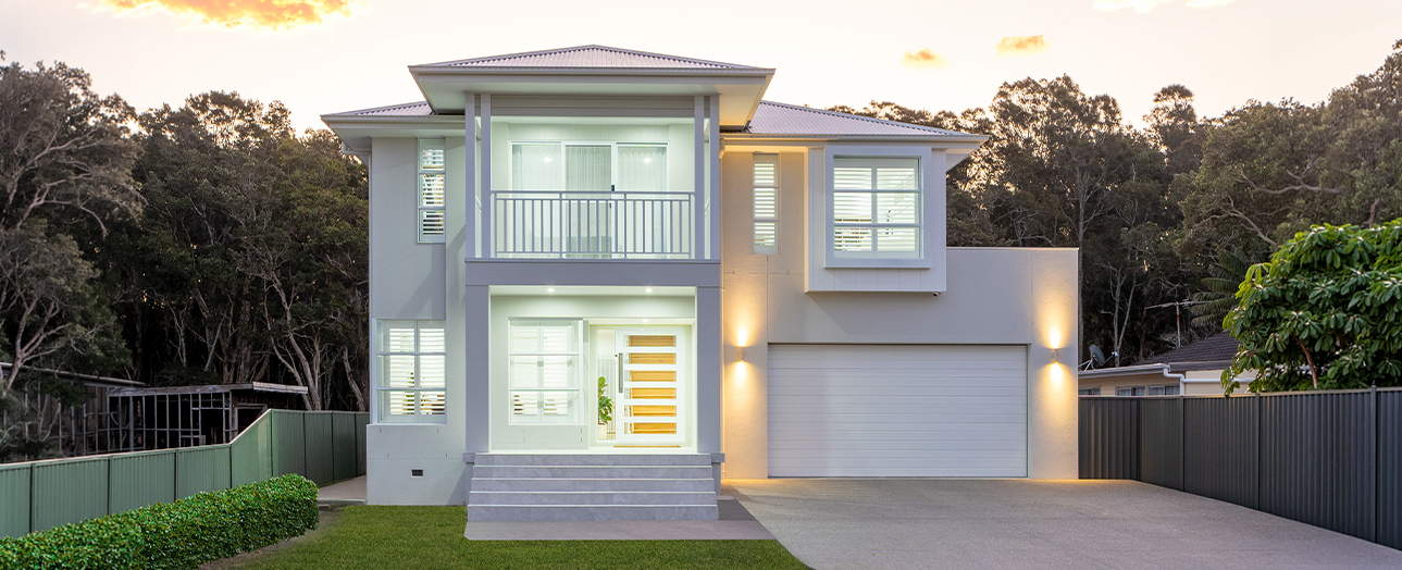 Modern two-story residential house in Noraville with white and gray exterior, featuring a double garage, upper-level balcony, large windows with shutters, and warm landscape lighting at dusk. Surrounded by native trees and manicured lawn.
