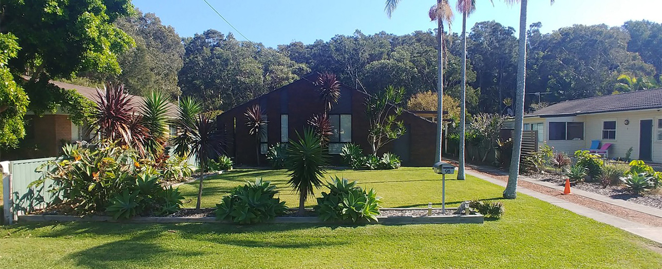 Noraville residential property before renovation, featuring a dark-colored single-story house with large windows, surrounded by mature palm trees and tropical vegetation, with a spacious green lawn and neighboring houses visible on either side, backed by dense bushland.