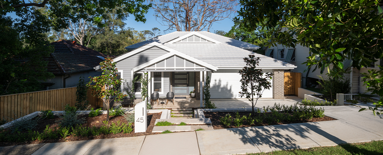 Modern white single-story house with pitched roof and brick accents at 45 Normanhurst. Front features covered porch with chairs, manicured landscaping with young trees and shrubs, concrete driveway, and wooden fence. Clear blue sky with mature trees visible in background.