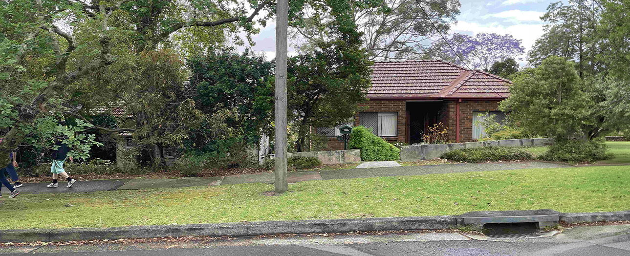 Single-story brick house with terracotta tile roof surrounded by mature trees and overgrown vegetation. A large lawn with fallen leaves and debris is in the foreground, with a person walking on the left side of the property. The house appears neglected with dense greenery obscuring parts of the structure.