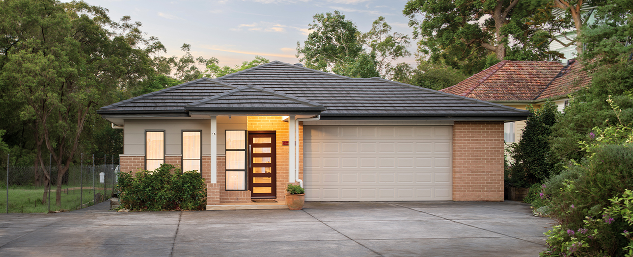 Modern single-story house with dark slate roof, cream and brick facade, illuminated front door with horizontal slats, large garage door, and manicured landscaping. Driveway in foreground with mature trees flanking the property against a twilight sky.
