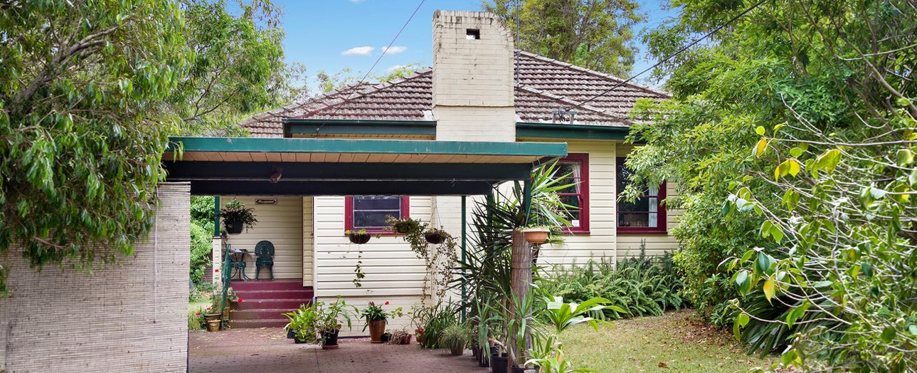 A single-story brick and weatherboard house with a dark tiled roof and central chimney, surrounded by overgrown vegetation and trees. The front entrance features a burgundy door beneath a turquoise awning, with hanging baskets and potted plants scattered around the porch. The property appears dated and somewhat neglected, with dense greenery encroaching on the sides and front yard.