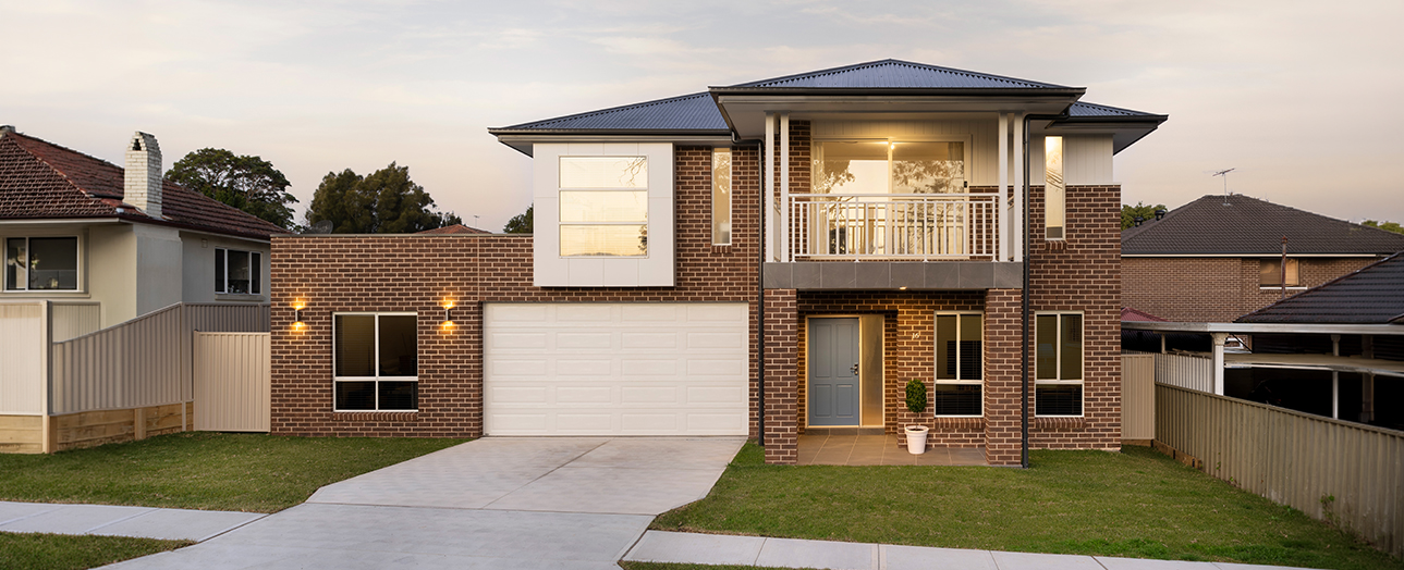 Modern two-story brick house in Parramatta with white garage door, blue metal roof, front balcony with white railings, illuminated windows at dusk, manicured lawn, concrete driveway, and neighboring houses visible on either side.