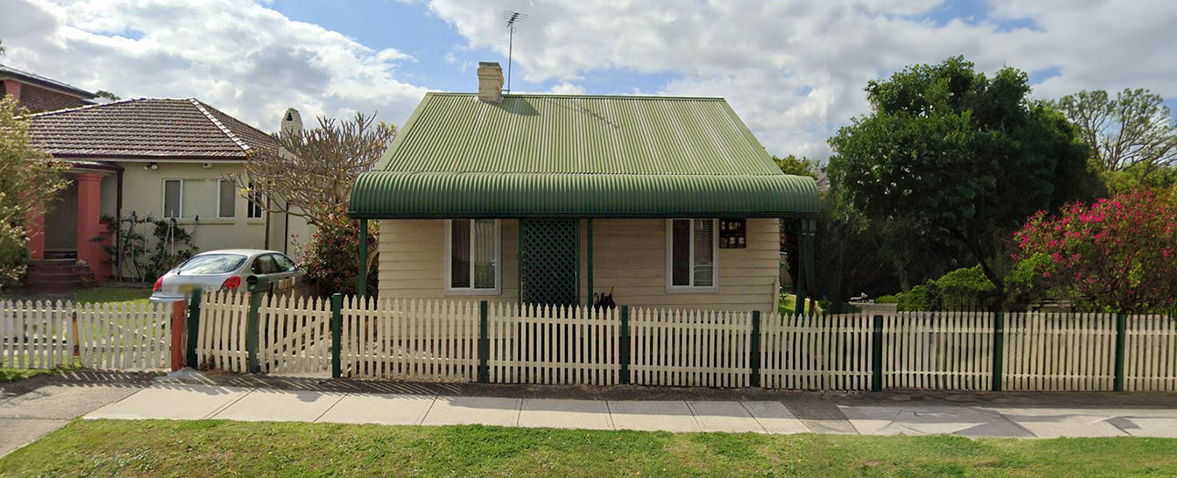 Parramatta heritage cottage with green corrugated iron roof and cream weatherboard walls, surrounded by white picket fence. Single-story house features dark green trim, front veranda, and is flanked by neighboring brick homes. Mature trees and flowering shrubs visible in yard and adjacent properties. Car parked on street.