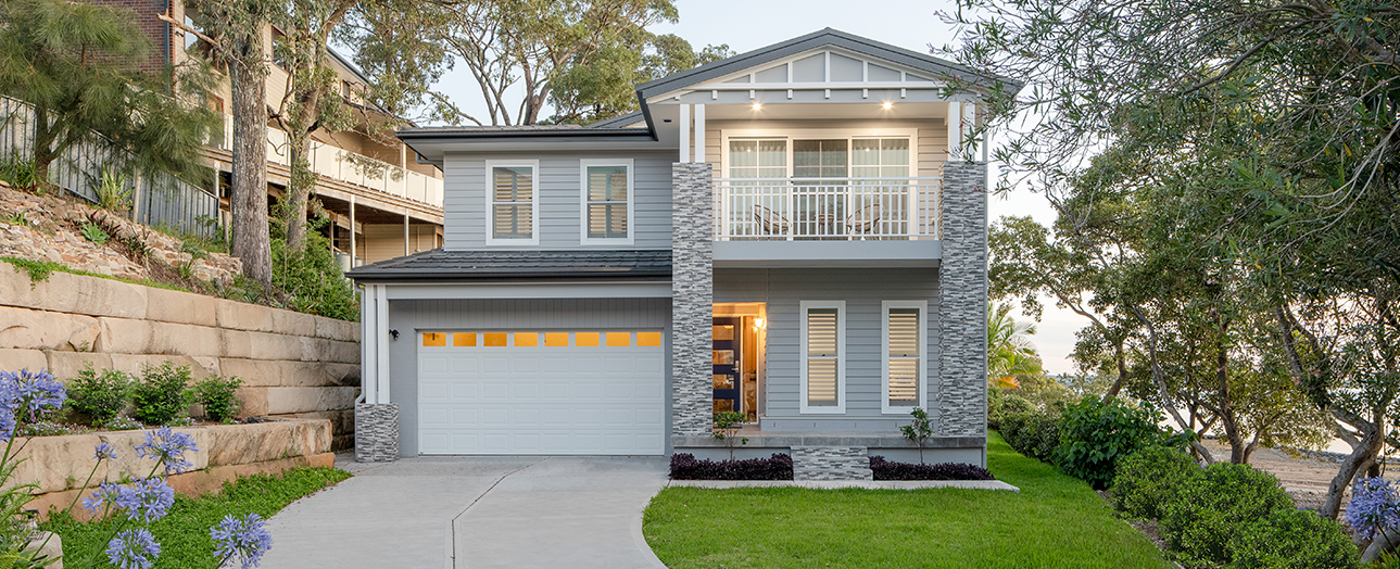 Modern two-story residential home in Point Clare with light gray and stone exterior, white garage door with yellow panels, second-floor balcony, manicured lawn, landscaping with blue flowering plants, concrete driveway, and surrounding trees.