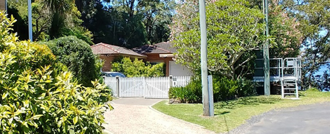Point Clare residential property before renovation, showing a single-story brick house with terracotta roof, white picket fence, gravel driveway, surrounding mature trees and landscaping, and water visible in the background.