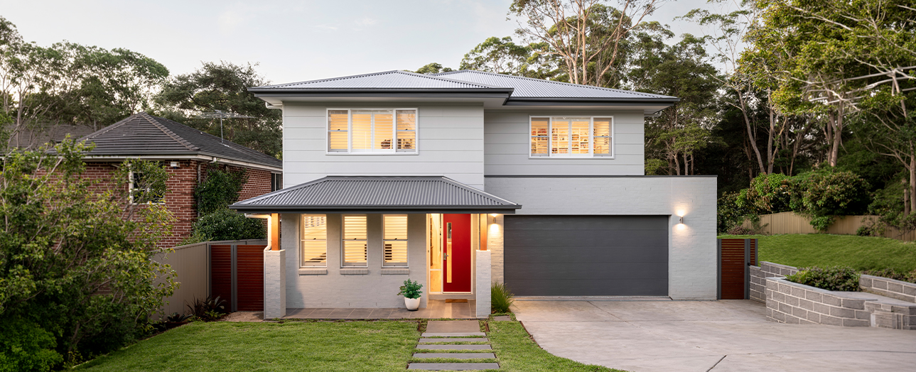 Modern two-story residential home in Pymble with white rendered facade, dark metal roof, large windows, red front door, and dark garage door. Property features manicured lawn, paved driveway, brick retaining wall, and mature trees surrounding the residence at dusk with interior lights glowing.