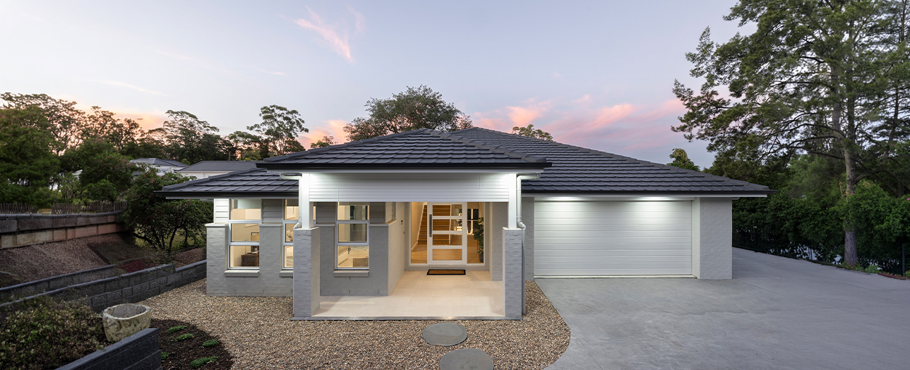 Modern single-story home with white exterior walls and dark hip roof, illuminated at dusk. The house features a central covered entry with glass doors, large windows, and an attached garage on the right. Gravel driveway and landscaping with trees surround the contemporary residence against a twilight sky with pink clouds.