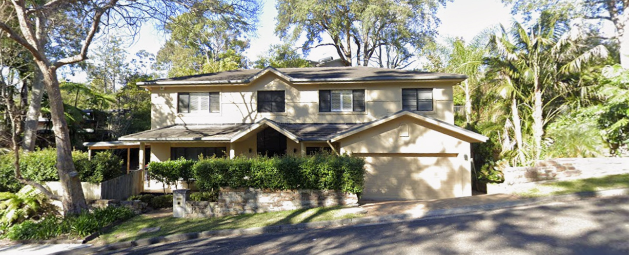 Two-story residential house with cream and white exterior, double garage, front porch, surrounded by mature trees and landscaping in a suburban setting before renovation.