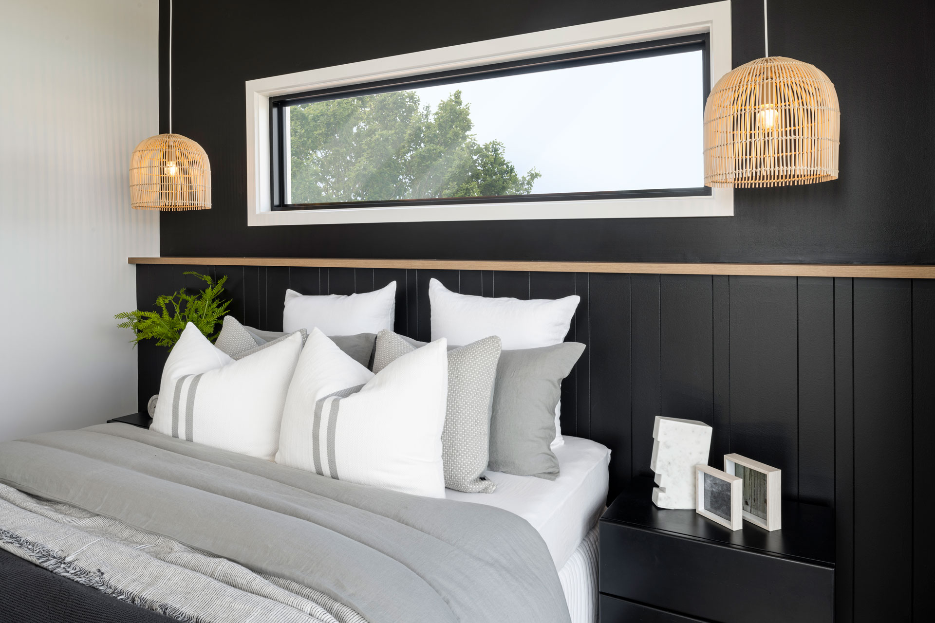 Modern bedroom with black paneled headboard and white bedding accented with gray and striped pillows. Two woven rattan pendant lights hang above the bed. A horizontal window with white frame overlooks trees. Wood shelf runs along the wall above the headboard. Potted green plant and framed photos sit on the nightstand.