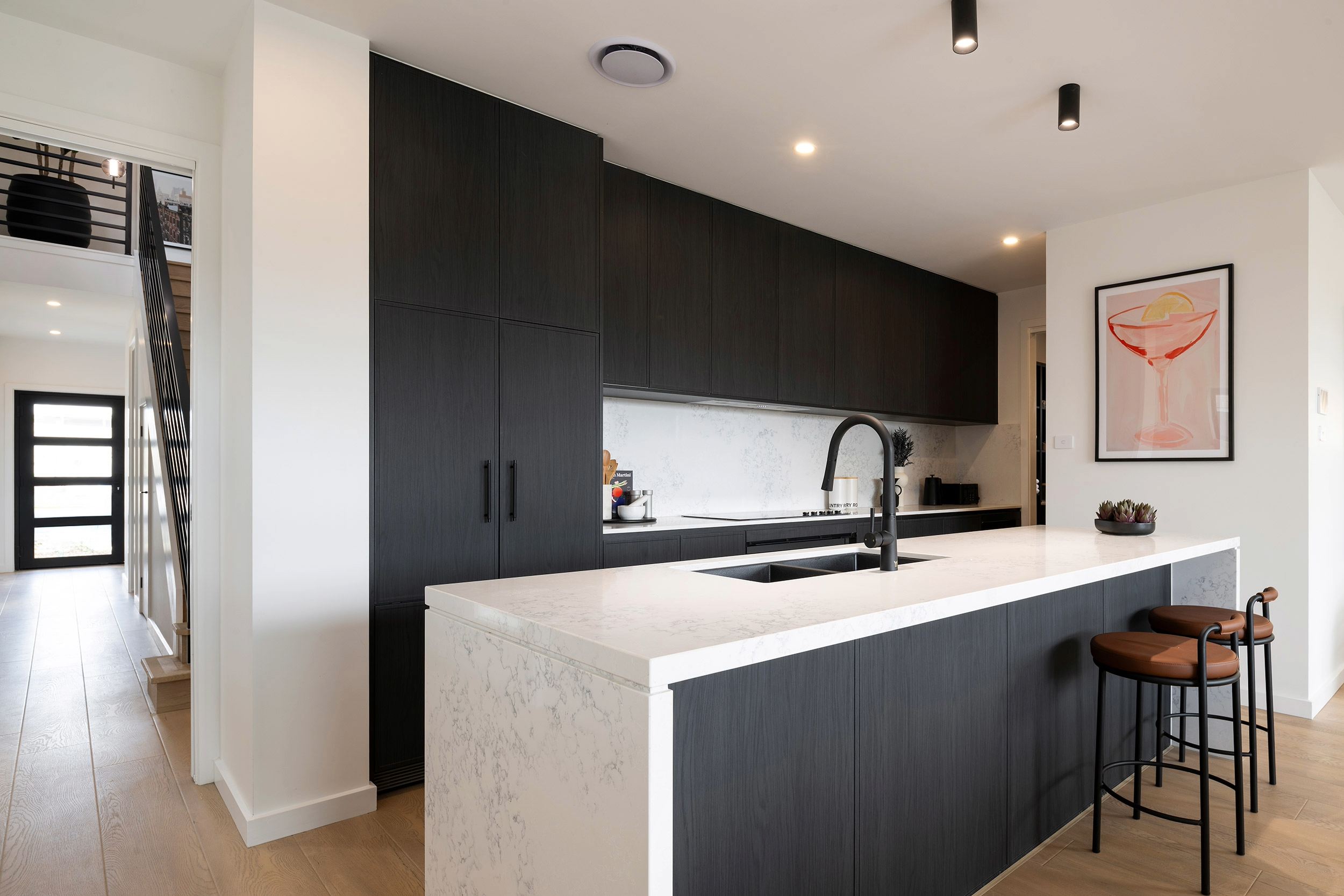 Modern kitchen with black cabinetry and white marble waterfall island countertop, featuring a black pull-down faucet and undermount sink. White subway tile backsplash, recessed lighting, and two brown leather bar stools at the island. Framed cocktail artwork on the white wall, black front door visible on the left, and light wood flooring throughout the open-plan space.