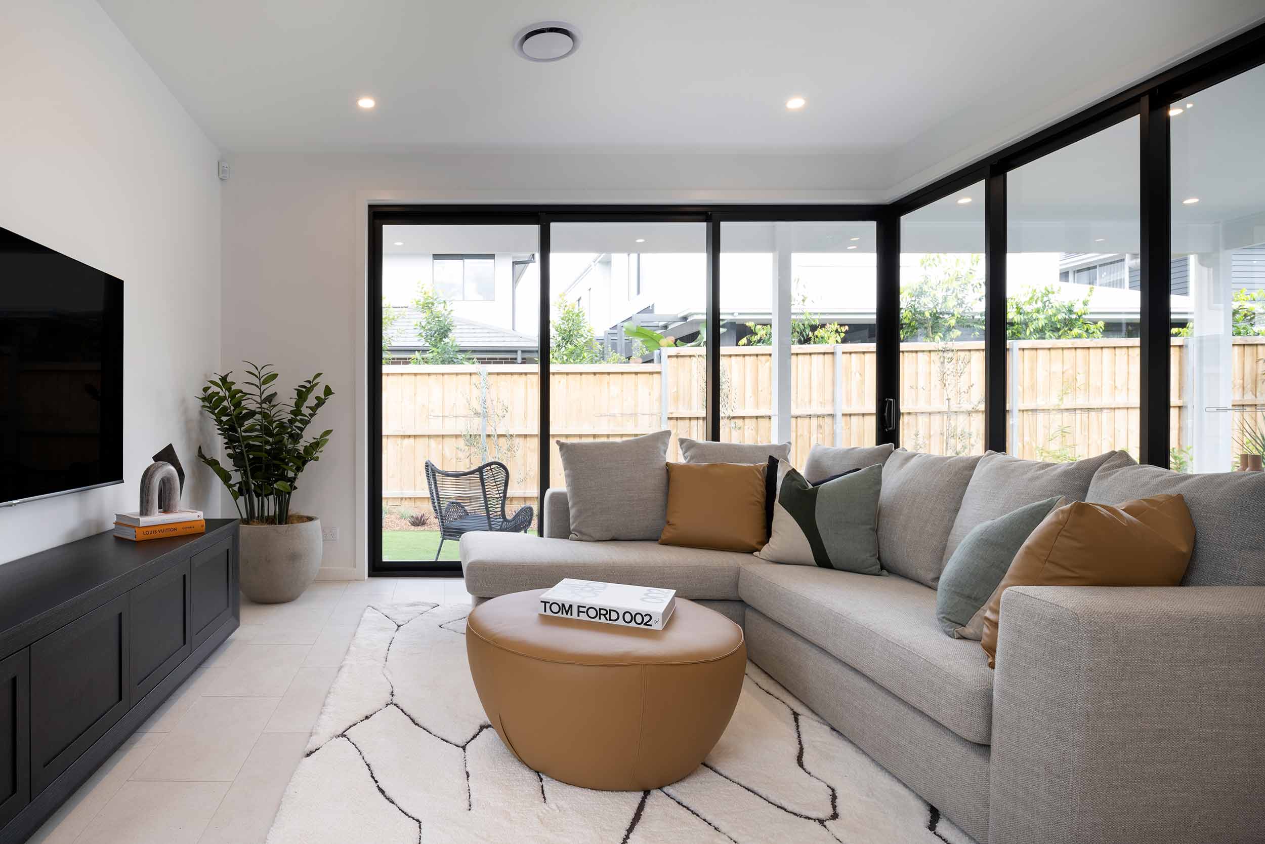 Modern living room with light gray sectional sofa, tan leather ottoman coffee table, and black-framed sliding glass doors opening to a wooden fence and backyard. A flat-screen TV mounted on a black media console sits on the left, with potted plants and decorative items nearby. The space features white walls, recessed lighting, and a geometric patterned area rug.