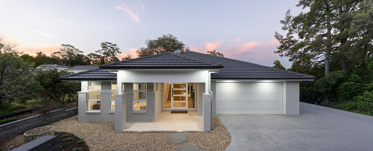 Modern single-story residential home with white and grey exterior, dark pitched roof, large windows, and attached garage, photographed at dusk with landscaped grounds and surrounding trees.