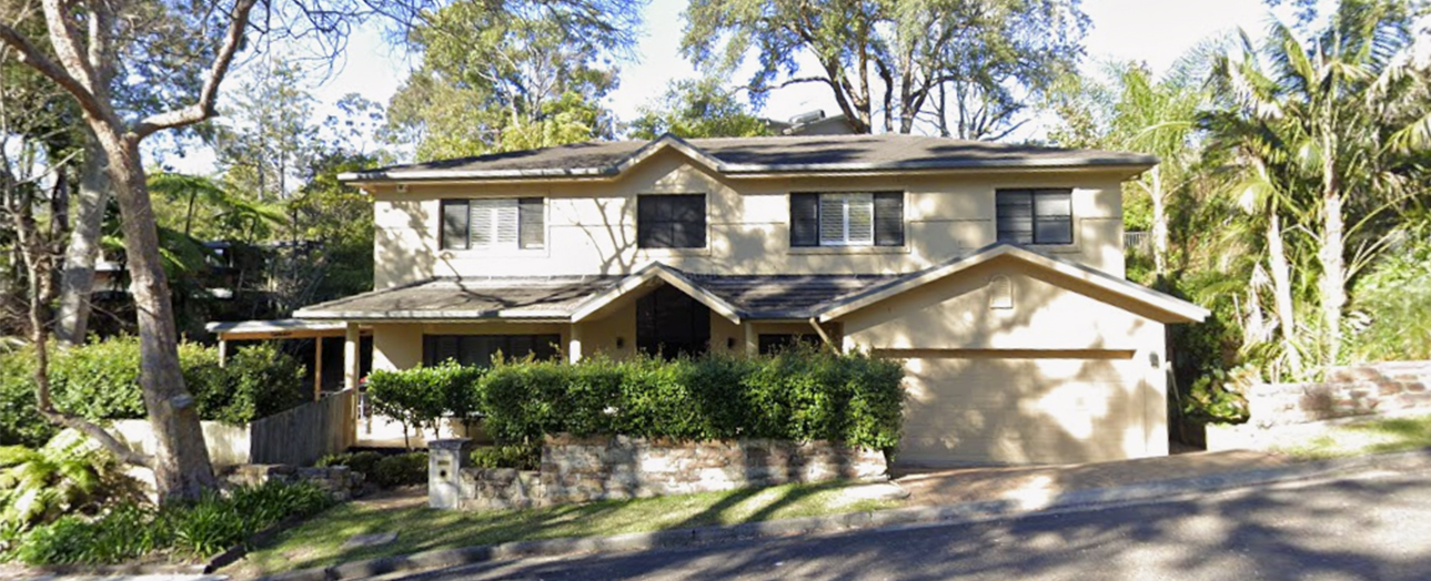 A two-story residential house with cream and tan exterior, featuring a pitched roof, front garage, and covered front porch. The property is surrounded by mature trees and green hedges, with a driveway leading to the garage. Clear blue sky visible above.