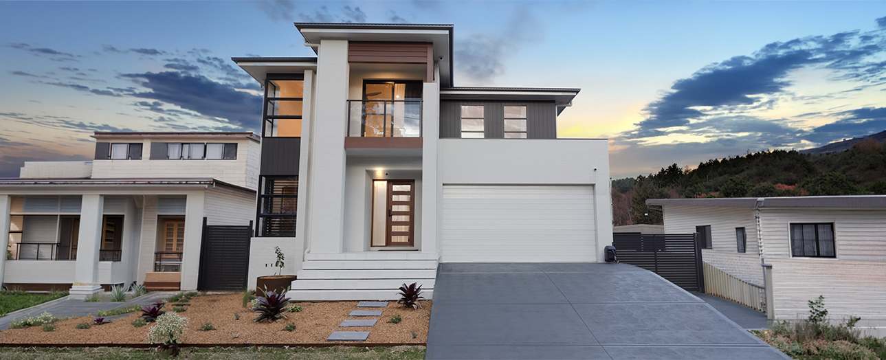 Modern two-story white and dark gray house with contemporary architecture, featuring a multi-level design, large windows, and a concrete driveway. Landscaped front yard with native plants and gravel. Neighboring houses visible on either side. Sunset sky with clouds in background and forested hills.