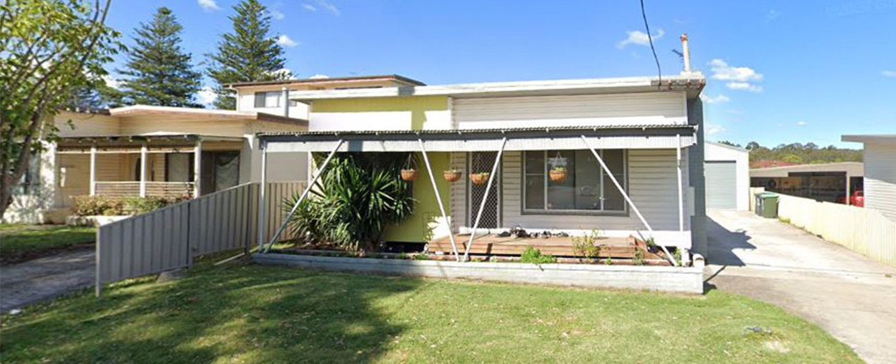Single-story residential house with yellow-green and white exterior, metal fencing, covered porch area, and grassy front yard before renovation.