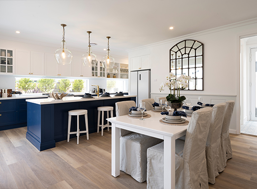 Modern kitchen and dining area with navy blue cabinetry, white countertops, pendant lighting, a white dining table with upholstered chairs, stainless steel appliances, and a decorative arched mirror on the wall.